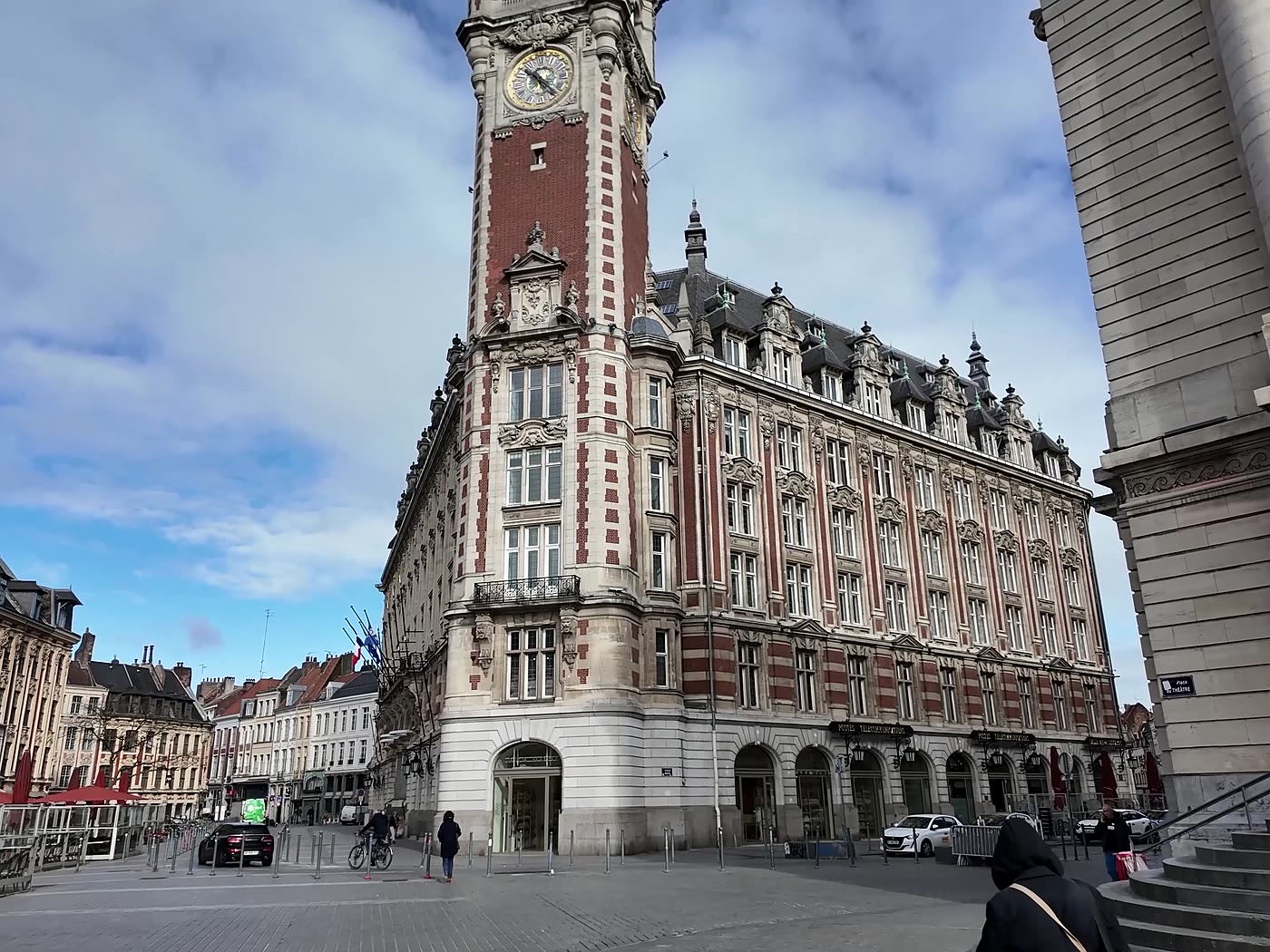 Beffroi à horloge et longue façade de brique rouge et de pierre blanche, sous un ciel nuageux.