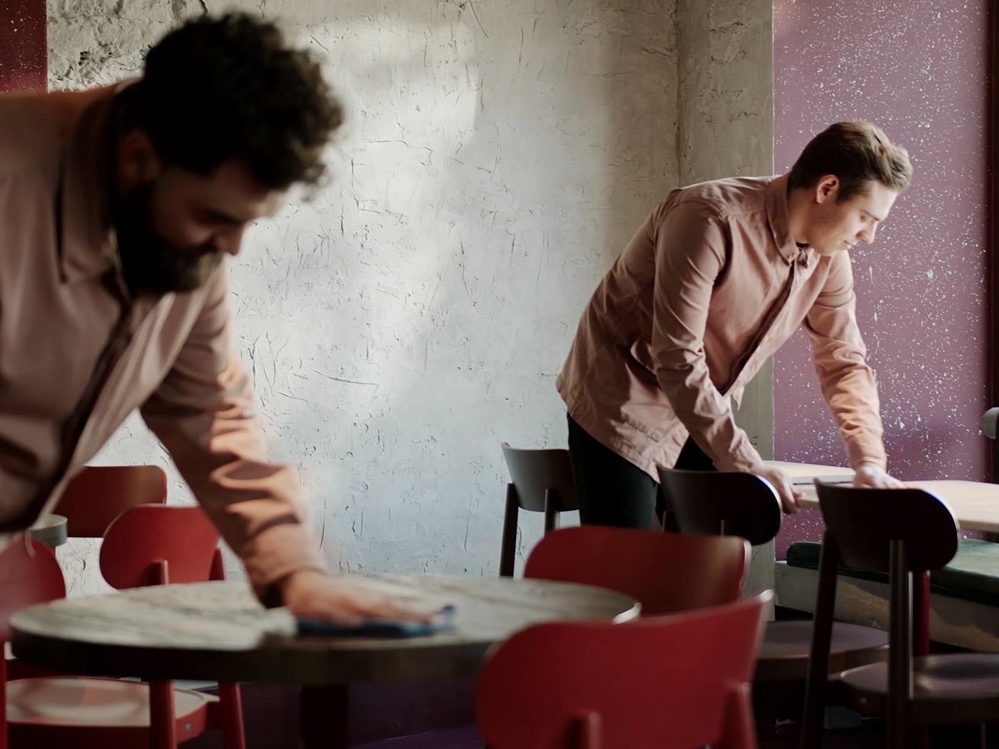Avant le service, deux membres de l’équipe de salle essuient et dressent les tables entourées de chaises rouges.