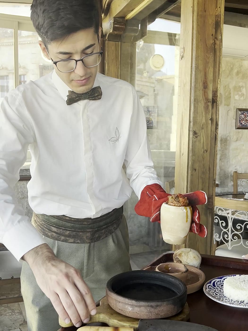 Un serveur en chemise blanche et nœud papillon présente à la table un plat mijoté dans un pot de terre.