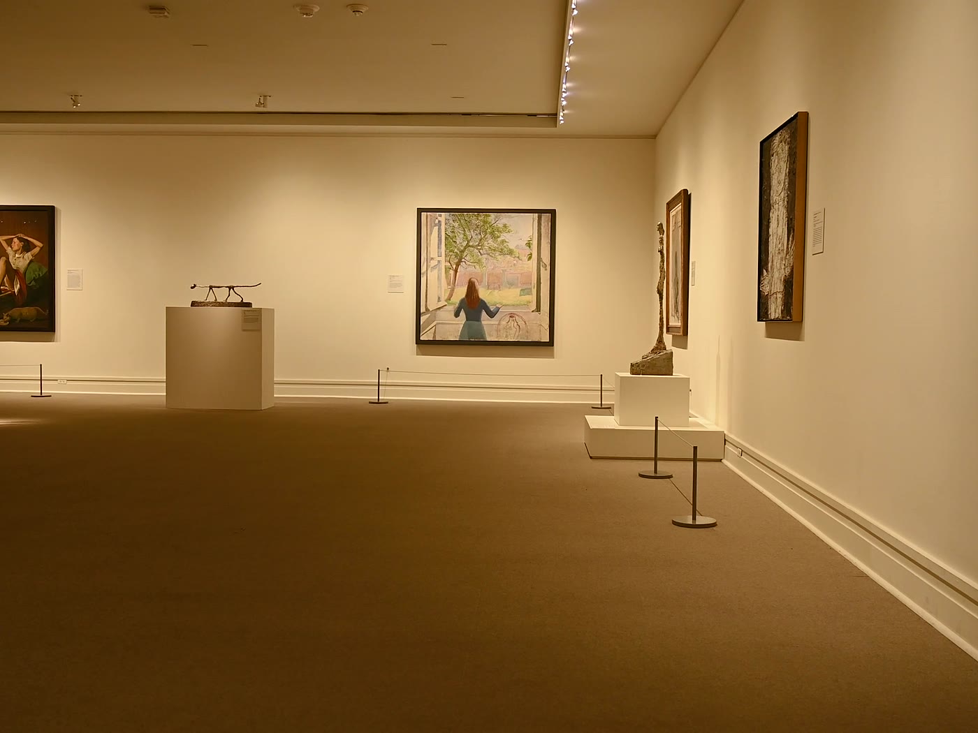 Salle de musée à la lumière chaude&nbsp;: tableaux encadrés sur des murs crème, deux sculptures filiformes sur socles blancs, cordons de mise à distance.