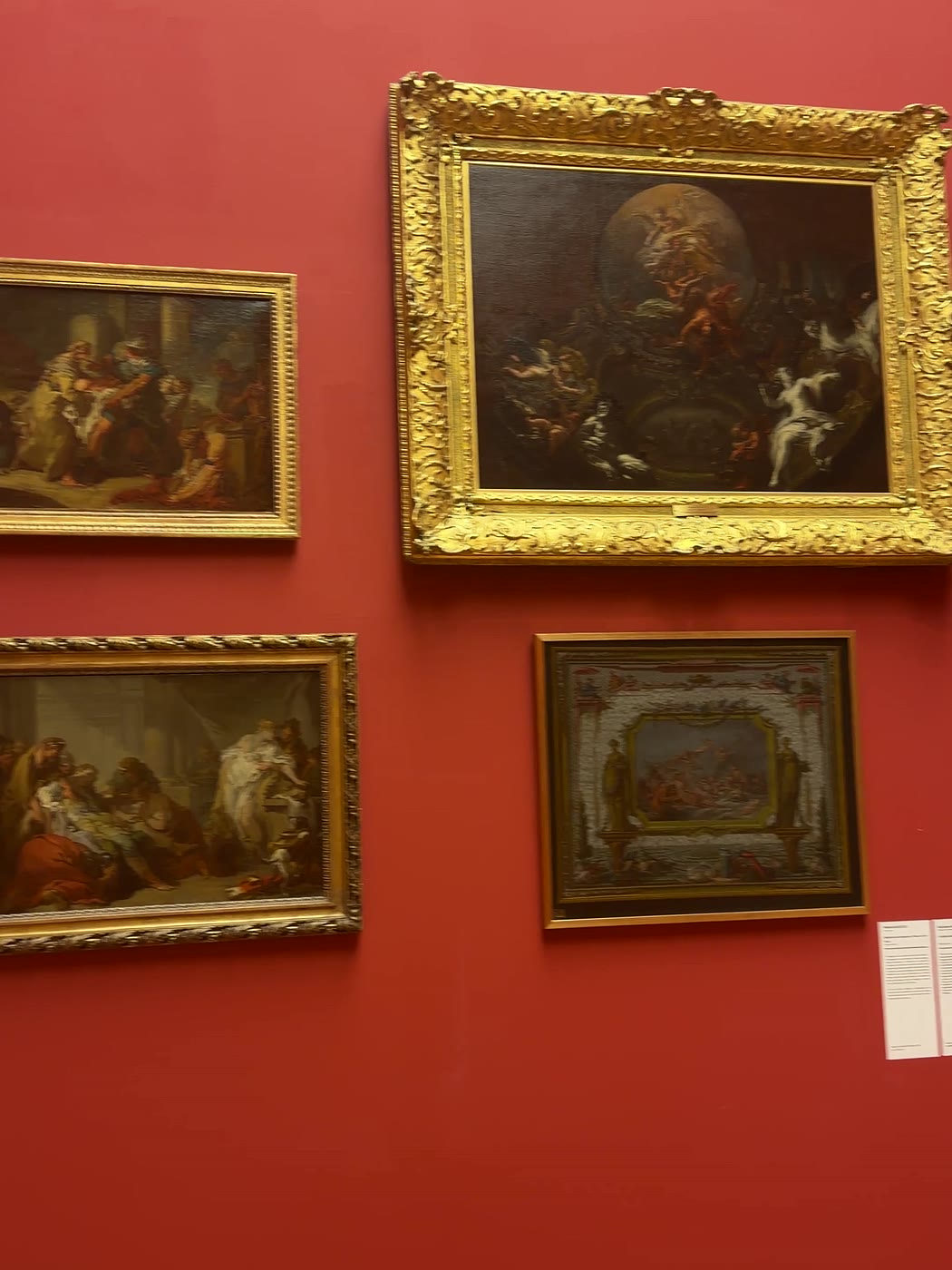 Mur rouge sombre d’un musée où sont accrochés quatre tableaux anciens dans de lourds cadres dorés.