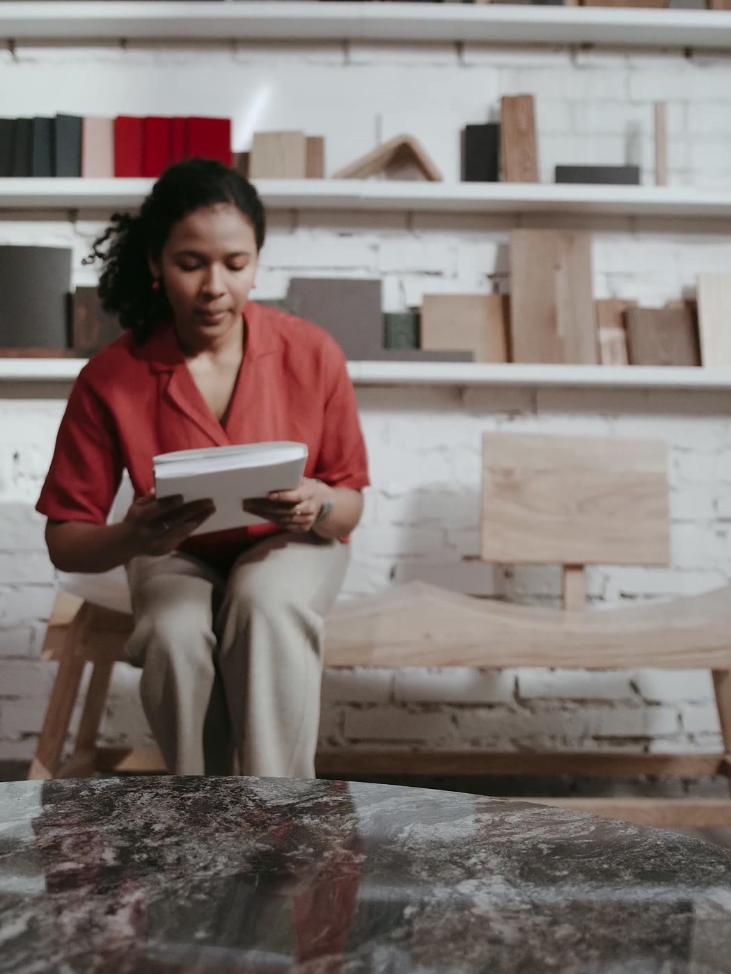 Une femme en chemise rouge feuillette un livre, assise sur un banc de bois devant des étagères d’échantillons et de livres.